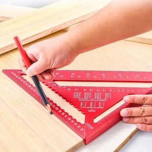 Puede incluir: Una escuadra de velocidad de aluminio roja y un lápiz marcan madera. La escuadra tiene medidas en pulgadas y tablas de conversión. El lápiz es negro con un extremo rojo.
