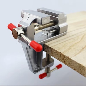 May include: A small, silver metal vise with red handles is clamped onto a piece of wood. The vise has a screw mechanism for tightening and loosening. The wood is light brown and has a rough, textured edge. This tool is used for holding objects in place.
