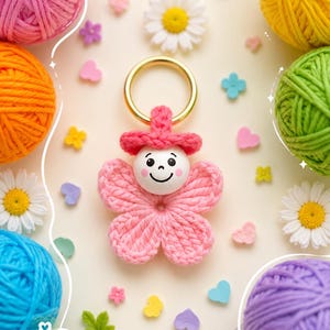 Può includere: Un portachiavi all'uncinetto con un corpo a forma di fiore rosa, un volto in perla di legno bianca con espressione sorridente e un cappellino all'uncinetto rosa. La figura è fissata a un anello in metallo dorato, circondata da gomitoli colorati.