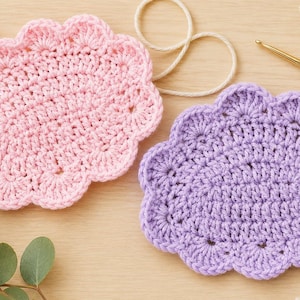 May include: Two oval-shaped crochet coasters, one in light pink and one in light purple, featuring a scalloped edge design. The coasters are crafted with a dense stitch pattern and are positioned alongside a gold-colored crochet hook.