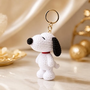 May include: A white crocheted dog keychain featuring a black nose, black ears, and a red collar. The figure is crafted with a textured yarn stitch and is attached to a gold-toned metal split ring by a short chain link for secure attachment.