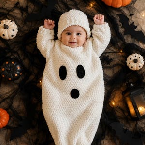 May include: A white crocheted baby sleeper with a matching beanie, featuring two black oval eyes and a black circular mouth to resemble a ghost. The garment has long sleeves and a textured knit pattern, designed for a baby to wear during Halloween.