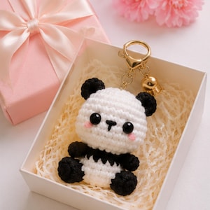 Op de afbeelding: Een gehaakte panda-sleutelhanger met een lichaam van wit en zwart garen, ronde zwarte ogen en roze geborduurde wangen. De panda is bevestigd aan een goudkleurige metalen karabijnhaak en een belletje, gepresenteerd in een witte geschenkdoos.