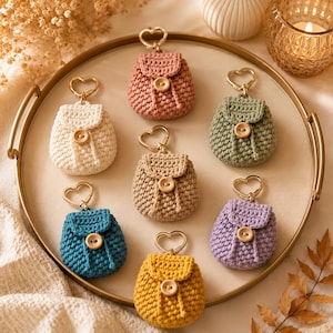 Può includere: Sette portachiavi a forma di zainetto realizzati all'uncinetto in vari colori tra cui rosa, bianco, verde, beige, blu, viola e giallo. Ogni piccola borsa presenta una patta con un bottone in legno e un anello dorato a forma di cuore.