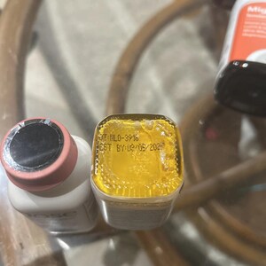 May include: A white bottle with a pink cap and a square-shaped bottle with a yellow liquid base. The square bottle features black text on the bottom reading LOT: HL0-3918 and BEST BY: 09/05/2024. The bottles are positioned upright on a reflective surface.