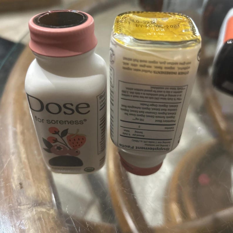 May include: Two small white plastic bottles of Dose for soreness supplement drink. One bottle stands upright with a pink cap and a label featuring a strawberry and blueberry graphic. The other bottle is inverted, showing the supplement facts label.