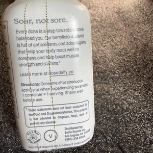 May include: A white plastic bottle with black text that reads: Soar, not sore. Every dose is a step towards a more balanced you. Our berrylicious blend is full of antioxidants and adaptogens that help your body react well to soreness and help boost muscle strength and stamina. Learn more at dosedaily.co. Directions: Consume after strenuous activity or when experiencing soreness. 1 container = 1 serving. Shake well before use.