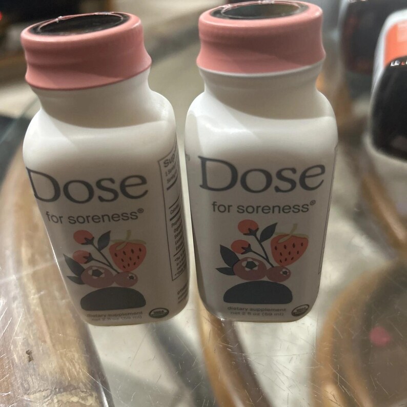 May include: Two white plastic bottles of Dose for soreness dietary supplement, each with a pink cap. The label features the text Dose for soreness and a graphic illustration of berries and leaves. Each bottle contains 2 fl oz (59 ml) of liquid.