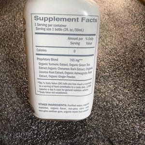 May include: A white plastic bottle featuring a Supplement Facts label. The text lists a 2 fl. oz. serving size and a 765 mg proprietary blend containing organic turmeric, green tea, cinnamon bark, licorice root, ashwagandha root, and ginger powder.