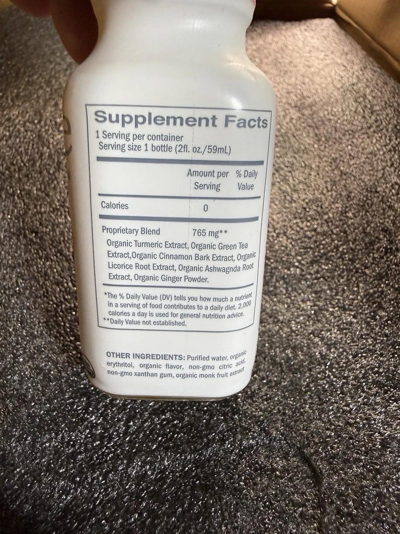 May include: A white plastic bottle featuring a Supplement Facts label. The text lists a 2 fl. oz. serving size and a 765 mg proprietary blend containing organic turmeric, green tea, cinnamon bark, licorice root, ashwagandha root, and ginger powder.