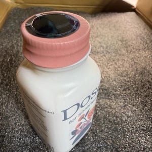 May include: A white plastic bottle with a light pink screw-on cap and a black circular seal on top. The label features the text "Dose" and "for soreness" along with a graphic illustration of orange and grey floral elements and text mentioning Curcumin.