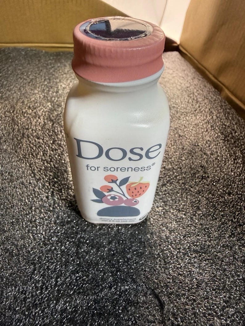 May include: A white plastic bottle with a pink cap containing a dietary supplement. The label features the text Dose for soreness and a graphic illustration of a strawberry and blueberries. The bottle has a net weight of 2.5 fl oz.
