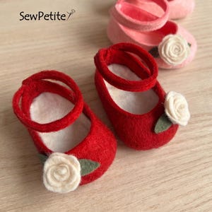 May include: Red felt baby shoes with white rose accents and ankle straps. The shoes are decorated with a white rose and green leaf. The text "SewPetite" is in the upper left corner. The shoes are on a light wood surface.