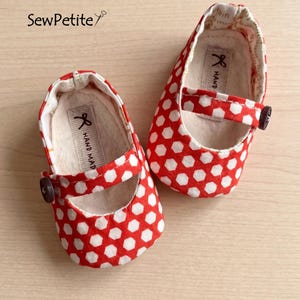 Puede incluir: Un par de zapatos de bebé de lunares rojos y blancos con una correa y cierre de botón. Los zapatos están hechos a mano y tienen un interior suave. El nombre de la marca "SewPetite" es visible en la esquina superior izquierda.