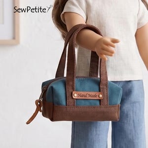 May include: A small, blue and brown handbag with brown handles and a leather tag that reads "Hand Made." The bag has a rectangular shape and a zipper closure. The bag is held by a doll.