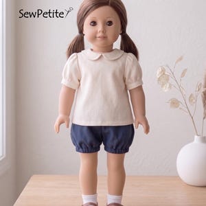May include: A doll wearing a cream-colored top with a Peter Pan collar and navy blue bloomers. The doll has brown hair in pigtails, brown shoes, and white socks. The text "SewPetite" is in the upper left corner.