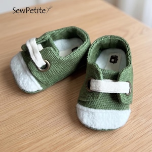 May include: A pair of small green canvas shoes with white accents. The shoes have white laces and a white toe cap. The text "SewPetite" is visible in the upper left corner. The shoes are on a light brown wooden surface.