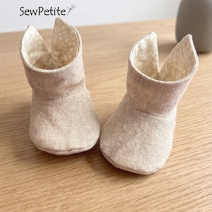 May include: Two beige fabric baby booties with a textured exterior and a patterned interior. The booties have a rounded toe and a tall, open top. The brand name "SewPetite" is visible in the upper left corner.