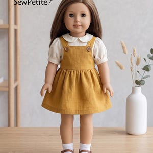Peut inclure: Une poupée vêtue d'une robe salopette jaune moutarde sur un chemisier blanc à col Claudine. La robe a deux boutons marron et une jupe évasée. La poupée a les cheveux bruns et porte des chaussures marron et des chaussettes blanches. Le texte "SewPetite" est visible.