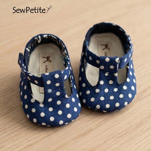 Op de afbeelding: Een paar babyschoentjes met een marineblauwe polka dot print. De schoentjes hebben een T-bandje en zijn handgemaakt. De tekst "SewPetite" is zichtbaar.
