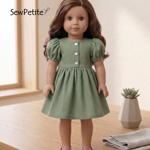 Könnte beinhalten: Eine Puppe in einem salbeigrünen Kleid mit kurzen, gepufften Ärmeln und einer Knopfleiste vorne. Das Kleid hat einen gerafften Rock und wird mit rosa Schuhen kombiniert. Die Puppe hat lange braune Haare und steht auf einem Holztisch. Der Text "SewPetite" ist sichtbar.