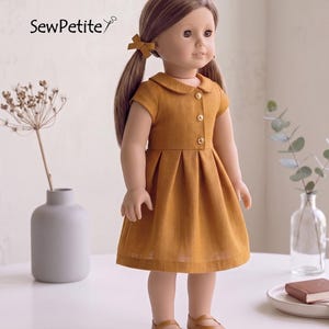 May include: A doll wearing a mustard-colored dress with a Peter Pan collar and button details. The doll has brown hair styled in pigtails with matching bows and is wearing brown shoes. The text "SewPetite" is in the upper left corner.