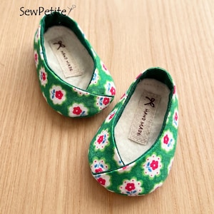 Könnte beinhalten: Ein Paar grüne Stoffpuppen-Schuhe mit einem Blumenmuster in Rot, Weiß und Blau. Die Schuhe haben eine grüne Einfassung und ein beiges Innenfutter. Jeder Schuh hat ein kleines Etikett mit der Aufschrift "HAND MADE".