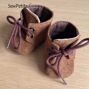 Könnte beinhalten: Ein Paar Miniatur-Stiefelchen aus braunem Wildleder mit passenden Schnürsenkeln. Die Stiefel haben Metallösen und ein weiches, strukturiertes Aussehen. Das Innenfutter ist dunkler braun, und die Stiefel sind für eine kleine Puppe oder ein Spielzeug gedacht.