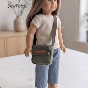 May include: A doll with long brown hair wearing a short-sleeved cream top, blue trousers, and beige shoes. The doll carries a small olive green crossbody bag with a brown strap and a leather-like tag labelled Hand Made.