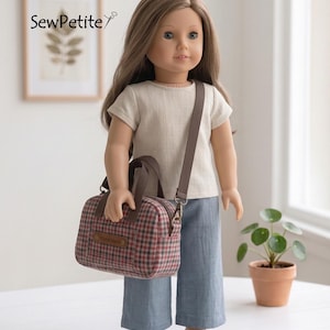 May include: A doll with long brown hair wearing a cream-colored short-sleeved shirt and blue trousers. The doll carries a small, rectangular, red and black plaid handbag with brown handles and a matching brown crossbody strap.