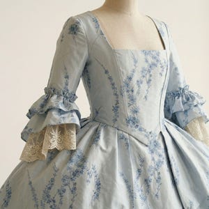 May include: A light blue dress featuring a dark blue floral pattern, a square neckline, and three-quarter length sleeves with double-tiered ruffles. The sleeve cuffs are finished with cream-coloured lace trim, and the bodice has a pointed waistline.