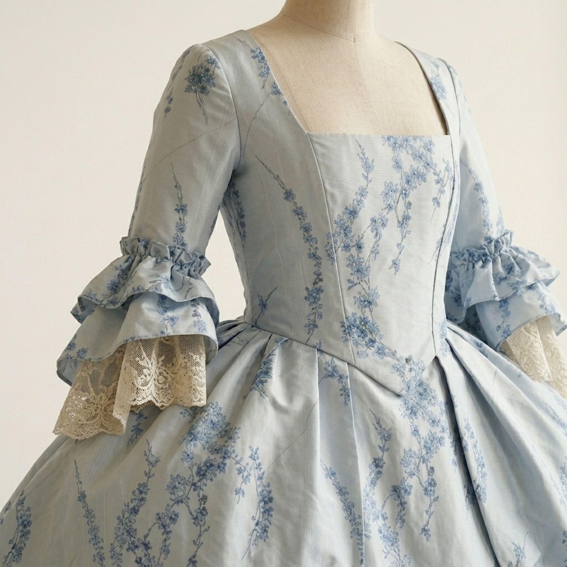 May include: A light blue dress featuring a dark blue floral pattern, a square neckline, and three-quarter length sleeves with double-tiered ruffles. The sleeve cuffs are finished with cream-coloured lace trim, and the bodice has a pointed waistline.