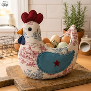 K&ouml;nnte beinhalten: Ein Stoffkorb in H&uuml;hnerform mit cremefarbenem Grund und einem rot-blauen Toile-Druck von Bauernhofszenen. Das Huhn hat einen roten Filzkamm, einen gelben Schnabel, einen blauen Kehllappen und einen blauen Filzfl&uuml;gel mit rotem Stern-Applikat.