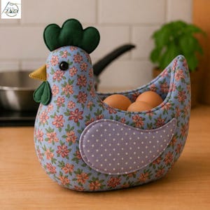 K&ouml;nnte beinhalten: Ein Eierhalter aus Stoff in Form eines Huhns mit hellblauem Grund und einem rosa-gr&uuml;nen Blumenmuster. Er hat einen dunkelgr&uuml;nen Kamm, einen gelben Schnabel, einen dunkelgr&uuml;nen Kehllappen und einen hellvioletten Fl&uuml;gel mit wei&szlig;en Punkten. Drei braune Eier liegen darin.
