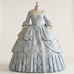May include: A light blue gown featuring a square neckline, three-quarter length sleeves with ruffled cuffs, and cream-coloured lace trim. The dress is adorned with a dark blue floral pattern, a fitted bodice, and a voluminous, pleated skirt.