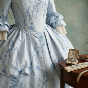 May include: A light blue gown with a dark blue floral pattern, featuring three-quarter length sleeves with ruffled cuffs and cream lace trim. A small open jewellery box containing a brooch and a folding fan are visible on a wooden surface.