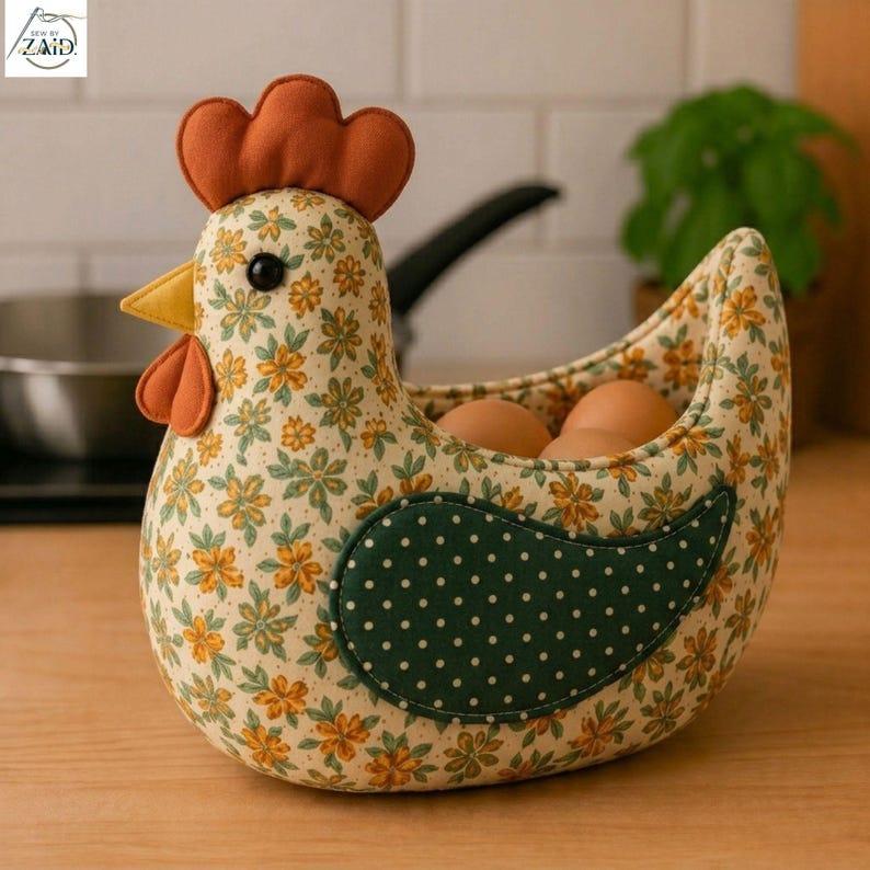 K&ouml;nnte beinhalten: Ein Eierhalter aus Stoff in Form eines Huhns mit cremefarbenem K&ouml;rper und gelb-gr&uuml;nem Blumenmuster. Er hat einen einfarbigen orangefarbenen Kamm und Kehllappen, einen gelben Schnabel und einen dunkelgr&uuml;nen Fl&uuml;gel mit wei&szlig;en Punkten. Drei braune Eier liegen darin.