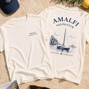 Flatlay of Natural Amalfi Sailing Club graphic tee with front chest logo and back sailboat artwork, styled with nautical summer details.
