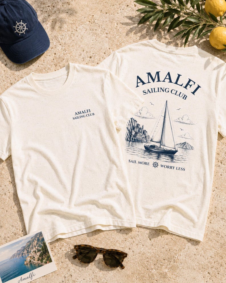 Flatlay of Natural Amalfi Sailing Club graphic tee with front chest logo and back sailboat artwork, styled with nautical summer details.