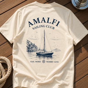 Natural Amalfi Sailing Club back print shirt flatlay on wooden yacht deck, featuring vintage sailboat artwork and Mediterranean coastal style.