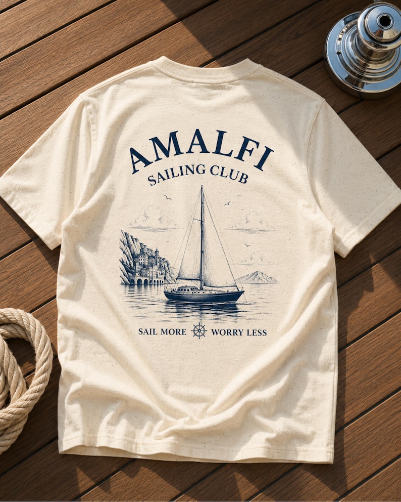 Natural Amalfi Sailing Club back print shirt flatlay on wooden yacht deck, featuring vintage sailboat artwork and Mediterranean coastal style.