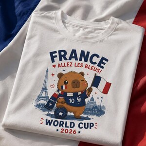 May include: A white t-shirt featuring a graphic of a brown capybara wearing a blue jersey with the number 10 and a scarf. The design includes the text FRANCE ALLEZ LES BLEUS! WORLD CUP 2026, the Eiffel Tower, a French flag, and a soccer ball.