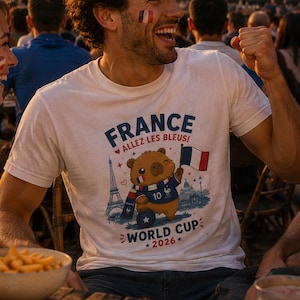 May include: A crowd watches a football match on a large screen. One person wears a white t-shirt with a bear graphic, the French flag, and the text "France, World Cup 2026". French flags are visible in the background, with fairy lights strung above.