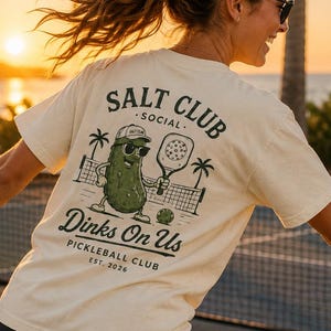 May include: A woman playing pickleball, wearing a cream-coloured t-shirt with a graphic of a pickle character and the text "SALT CLUB SOCIAL Dinks On Us PICKLEBALL CLUB EST. 2026". A yellow ball is in the air.