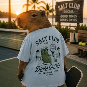 May include: A light blue t-shirt featuring a graphic of a pickle wearing sunglasses and a cap, holding a pickleball paddle. The text reads SALT CLUB SOCIAL, Dinks On Us, PICKLEBALL CLUB, EST. 2026. The design includes palm trees and a net.