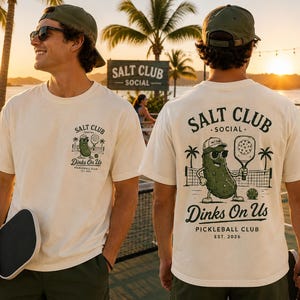 May include: An off-white t-shirt featuring a graphic of a pickle wearing sunglasses and a cap, holding a pickleball paddle. The text reads SALT CLUB SOCIAL, Dinks On Us, PICKLEBALL CLUB, EST. 2026. The design appears on the front chest and back.
