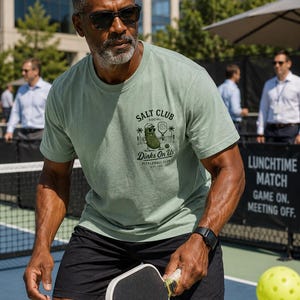 May include: A sage green short-sleeve crew neck t-shirt featuring a graphic on the chest. The print displays a cartoon pickle wearing sunglasses, holding a pickleball paddle, with the text SALT CLUB, SOCIAL, Dinks On Us, and PICKLEBALL CLUB EST. 2024.