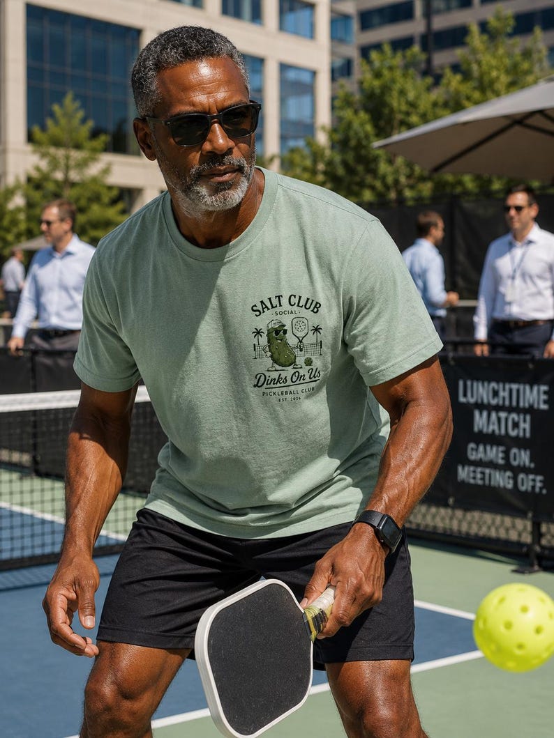 May include: A sage green short-sleeve crew neck t-shirt featuring a graphic on the chest. The print displays a cartoon pickle wearing sunglasses, holding a pickleball paddle, with the text SALT CLUB, SOCIAL, Dinks On Us, and PICKLEBALL CLUB EST. 2024.