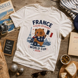 May include: A white short-sleeve t-shirt featuring a graphic of a brown capybara wearing a blue soccer jersey with the number 10, a scarf, and holding a French flag. The text reads FRANCE ALLEZ LES BLEUS! WORLD CUP 2026 with an Eiffel Tower illustration.
