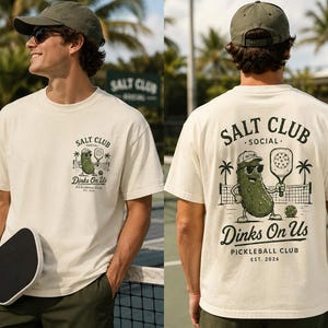 May include: An off-white short-sleeve t-shirt featuring a graphic of a pickleball-playing pickle wearing sunglasses and a cap. The front chest displays a small logo, while the back features a larger version with the text Salt Club Social Dinks On Us Pickleball Club Est. 2026.